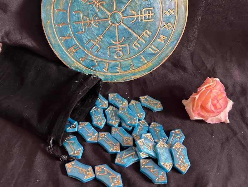 Op de afbeelding: Een set blauwe en gouden runenstenen met gouden belettering op elke steen. De stenen zijn verspreid over een zwart oppervlak. Een blauw en gouden rond houten bord met runen is op de achtergrond.