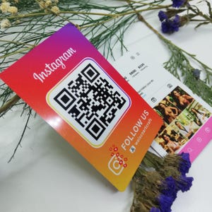 Op de afbeelding: Een visitekaartje met een rode, oranje en gele gradiënt, met een grote QR-code en het woord "Instagram" in witte letters. De kaart bevat ook de tekst "Follow Us" en een Instagram-logo. Een andere kaart toont een Instagram-profiel.