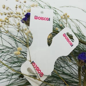 Op de afbeelding: Twee witte productlabels met een gebogen ontwerp en een gat om op te hangen. De labels hebben het woord "Woiscol" in roze gedrukt. De labels zijn omgeven door gedroogde bloemen en groen.