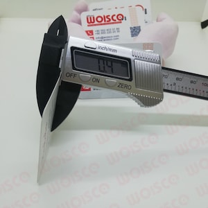 May include: A digital caliper measuring a white card. The caliper displays a measurement of 0.4 inches. The tool is silver with a black measuring arm and is set against a white background. The card has the text "WOISCO".