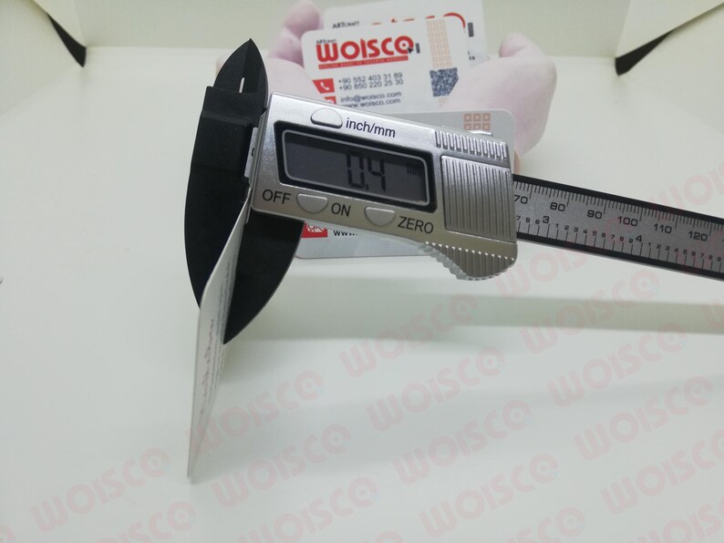 May include: A digital caliper measuring a white card. The caliper displays a measurement of 0.4 inches. The tool is silver with a black measuring arm and is set against a white background. The card has the text "WOISCO".