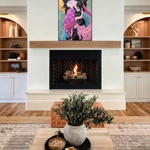 May include: A living room with a fireplace and artwork. The artwork features a woman surrounded by black cats. A wooden mantel sits above the fireplace. A coffee table with a tray, bowl, and plant is in front of the fireplace.