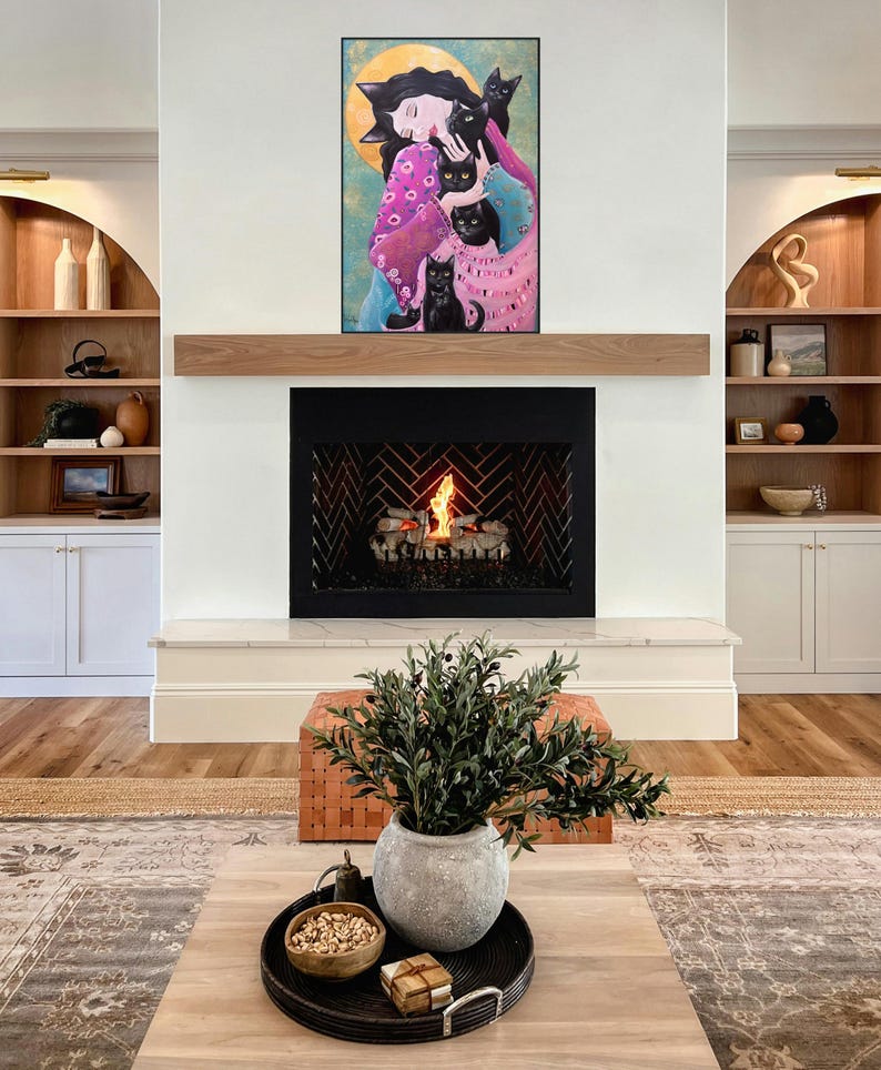 May include: A living room with a fireplace and artwork. The artwork features a woman surrounded by black cats. A wooden mantel sits above the fireplace. A coffee table with a tray, bowl, and plant is in front of the fireplace.