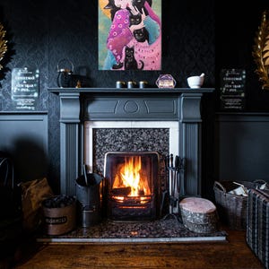 May include: A colorful painting of a woman with black cats hangs above a fireplace with a lit fire. The painting features a woman in a pink and blue dress, surrounded by black cats. The fireplace is dark gray with a marble hearth.