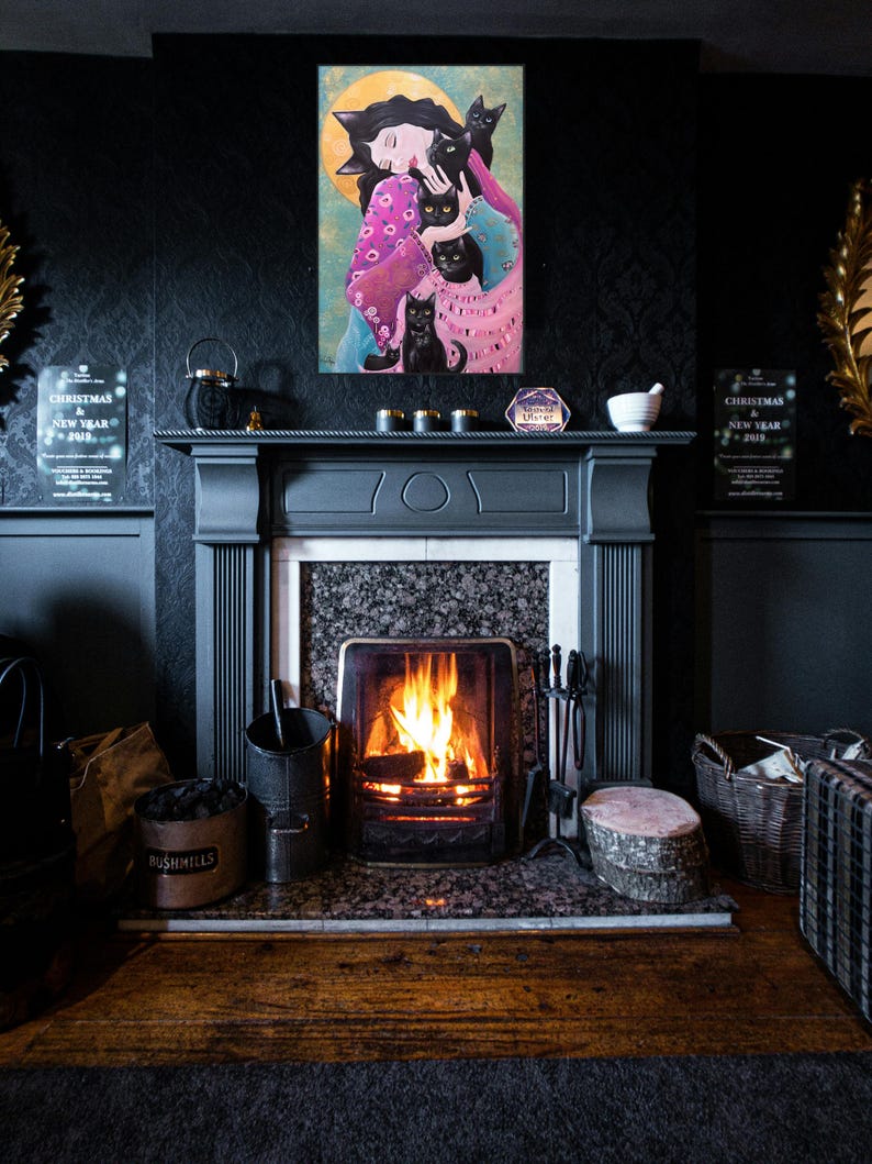 May include: A colorful painting of a woman with black cats hangs above a fireplace with a lit fire. The painting features a woman in a pink and blue dress, surrounded by black cats. The fireplace is dark gray with a marble hearth.