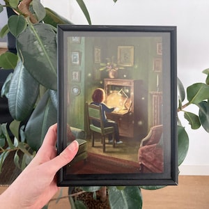 Op de afbeelding: Ingelijste kunstdruk met een persoon aan een bureau verlicht door een lamp. De scène speelt zich af in een kamer met een vintage esthetiek, met een bureau, stoel en ander meubilair. Het kunstwerk heeft een warm kleurenpalet met een focus op het licht.