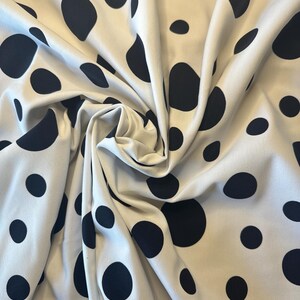 May include: Cream-colored fabric with a repeating pattern of large, irregularly shaped black polka dots. The fabric is draped and folded, creating shadows and texture. The polka dots vary in size and are scattered across the surface.