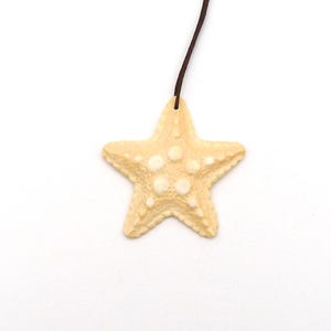 May include: A light beige starfish pendant with white raised dots, suspended by a thin brown cord. The starfish has five points and a textured surface, set against a white background.