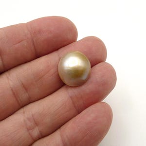May include: A lustrous, golden-colored pearl with a smooth, rounded surface. The pearl is held in a hand against a white background, showcasing its natural iridescence and warm tones. This gemstone is a classic element in jewelry design.