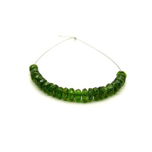 May include: A strand of small, faceted, forest green gemstone beads arranged in a curved line. Each bead features a circular, flattened shape with multiple reflective surfaces, strung together on a thin, light-colored flexible wire.