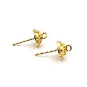 May include: Two gold-colored earring posts with a textured, bell-shaped design. Each post has a small loop at the top and a straight, slender post for insertion. The earrings are set against a white background.