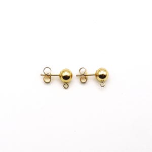 May include: A pair of gold-toned stud earrings featuring a smooth, spherical ball design. Each earring includes a post back and a small, circular jump ring loop attached to the bottom of the sphere, allowing for the addition of charms or dangles.