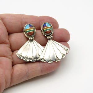 May include: A pair of silver earrings featuring a fan-shaped base with a scalloped edge. Each earring has an oval top section inlaid with segments of blue, white, green, and reddish-brown stone, surrounded by a twisted silver rope border.