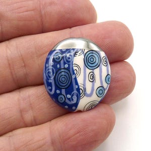May include: A round, blue and white ceramic button with a silver metal top. The button features a design of blue swirls and concentric circles on a white background. The button is held in a hand.