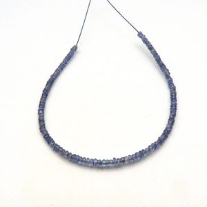 May include: A single strand of small, faceted, periwinkle blue gemstone beads arranged in a curved line. The beads are uniform in size and shape, featuring a translucent quality and a subtle sparkle from the light reflecting off their cut surfaces.