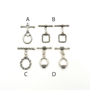 May include: A collection of silver-toned metal toggle clasps for jewelry making, labeled A, B, C, and D. The set includes various designs featuring heart-shaped, square, and circular rings with textured bars and loops for secure fastening.