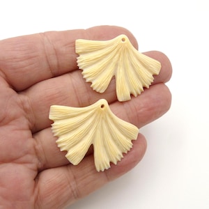 May include: Two cream-colored, fan-shaped pendants with a hole at the top for stringing. The pendants have a textured, ribbed design and a scalloped edge. They are displayed on a light background.