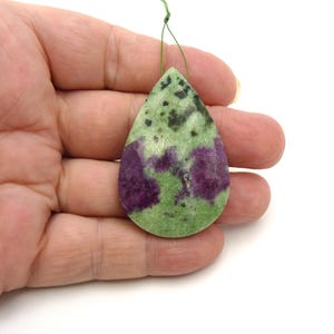 May include: A teardrop-shaped pendant made of zoisite stone featuring a mottled pattern of light green and dark green with prominent patches of deep magenta. The stone has a smooth, polished surface and a small hole at the top for suspension.