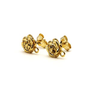 May include: A pair of gold-colored stud earrings. Each earring features a detailed rose design with a textured surface and a small loop at the bottom. The earrings are set against a white background.
