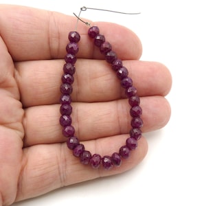 May include: A strand of faceted, deep red ruby beads. The beads are strung on a wire, forming a teardrop shape. The beads are held in a hand against a white background. The beads have a polished, reflective surface.