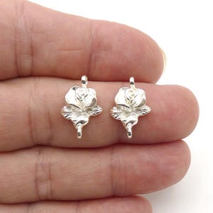 May include: Two silver-colored metal rose charms, each featuring a detailed floral bloom with leaves and a small loop at both the top and bottom for attachment. The charms have a polished, reflective surface and a symmetrical, three-dimensional design.