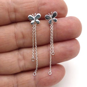 May include: A pair of silver butterfly earrings. Each earring features a butterfly stud with dangling chains. The chains have small, round charms. The earrings are displayed against a white background.
