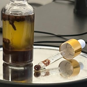 May include: A clear glass dropper bottle containing a yellow liquid with dark brown sediment and whole cloves at the bottom. A separate glass dropper with a gold-colored collar and a white rubber bulb lies horizontally next to the bottle.