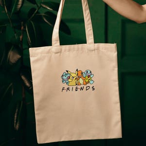 Pode incluir: Bolsa tote bege com um gr&aacute;fico colorido de personagens de desenhos animados e a palavra "FRIENDS" em preto. A bolsa tem duas al&ccedil;as e &eacute; segurada contra um fundo verde. O gr&aacute;fico inclui um personagem amarelo, um personagem laranja e dois personagens azuis.