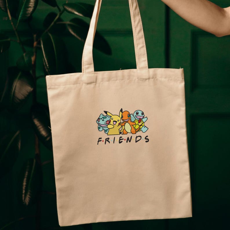 Pode incluir: Bolsa tote bege com um gr&aacute;fico colorido de personagens de desenhos animados e a palavra "FRIENDS" em preto. A bolsa tem duas al&ccedil;as e &eacute; segurada contra um fundo verde. O gr&aacute;fico inclui um personagem amarelo, um personagem laranja e dois personagens azuis.