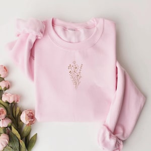 Puede incluir: Una sudadera rosa claro con un dise&ntilde;o floral bordado en color marr&oacute;n claro. La sudadera de cuello redondo est&aacute; cuidadosamente doblada, con las mangas metidas. El dise&ntilde;o floral est&aacute; centrado en el pecho.