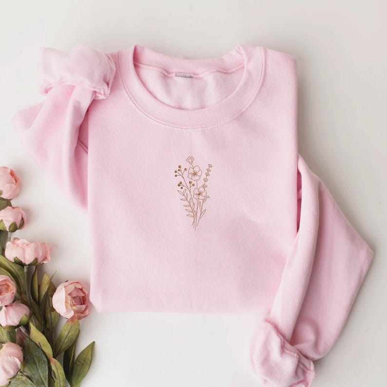 Puede incluir: Una sudadera rosa claro con un dise&ntilde;o floral bordado en color marr&oacute;n claro. La sudadera de cuello redondo est&aacute; cuidadosamente doblada, con las mangas metidas. El dise&ntilde;o floral est&aacute; centrado en el pecho.