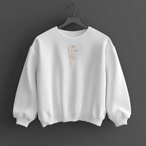 Puede incluir: Una sudadera blanca con cuello redondo y mangas largas. Presenta un peque&ntilde;o dise&ntilde;o floral bordado en color marr&oacute;n claro en la parte delantera. La sudadera est&aacute; colgada de una percha negra sobre un fondo gris oscuro.