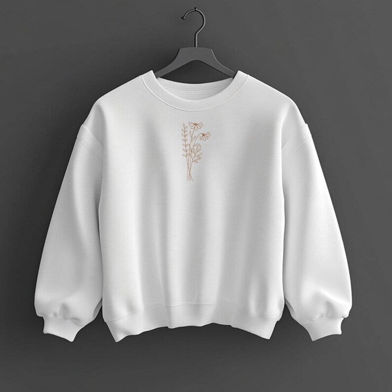 Puede incluir: Una sudadera blanca con cuello redondo y mangas largas. Presenta un peque&ntilde;o dise&ntilde;o floral bordado en color marr&oacute;n claro en la parte delantera. La sudadera est&aacute; colgada de una percha negra sobre un fondo gris oscuro.
