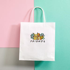 Pode incluir: Saco de papel branco com um gr&aacute;fico de desenho animado de personagens e a palavra "FRIENDS" em preto. O saco tem al&ccedil;as brancas e est&aacute; sobre um fundo rosa e turquesa.