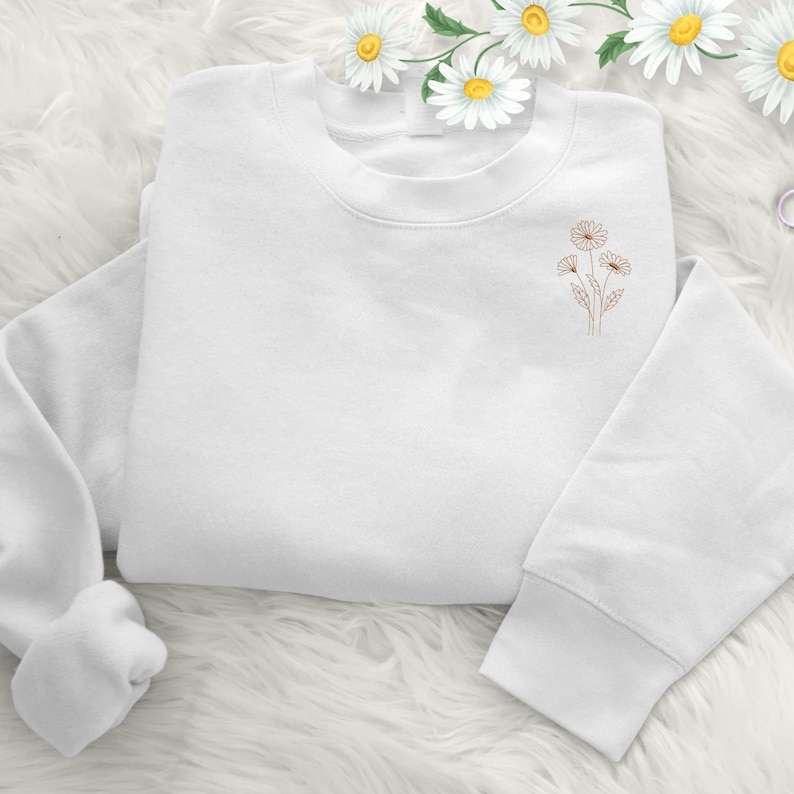 Puede incluir: Una sudadera blanca con cuello redondo y mangas largas. Presenta un peque&ntilde;o dise&ntilde;o bordado de tres margaritas en marr&oacute;n claro en la parte superior izquierda del pecho. La prenda se muestra sobre una superficie blanca y esponjosa.
