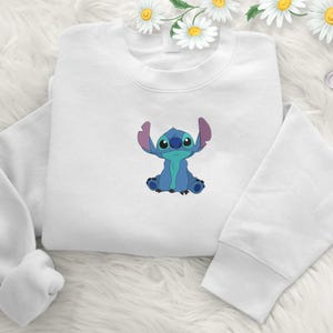 May include: White sweatshirt with a blue cartoon character embroidered on the front. The character has large ears and is sitting with its arms raised. The sweatshirt has long sleeves and a crew neck.