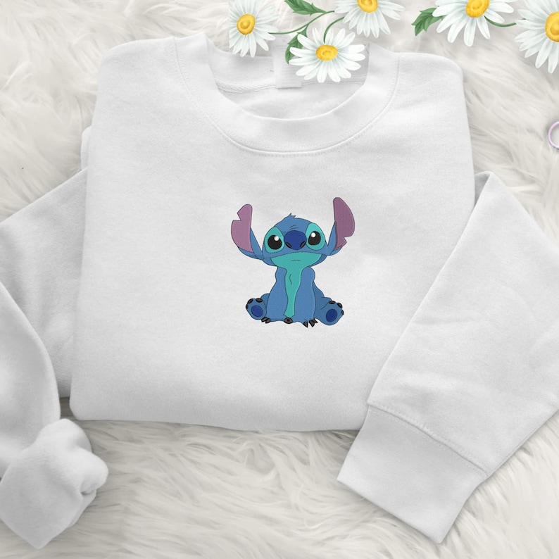May include: White sweatshirt with a blue cartoon character embroidered on the front. The character has large ears and is sitting with its arms raised. The sweatshirt has long sleeves and a crew neck.