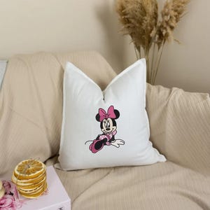 May include: White throw pillow featuring an embroidered Minnie Mouse design. Minnie Mouse is depicted in black, pink, and white, with a large pink bow. The pillow is on a beige sofa with a beige throw blanket.