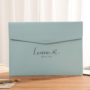 Puede incluir: Carpeta de documentos azul claro con cierre de solapa. La carpeta tiene el nombre "Lonin" y la fecha "April 13, 2026" impresos en negro. Un bolígrafo dorado y un portátil están sobre el escritorio de madera.