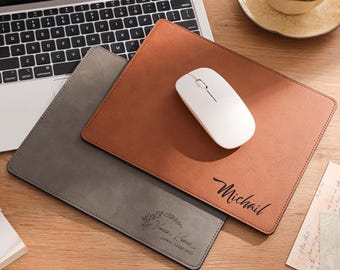 Op de afbeelding: Een laptop, twee leren muismatten, een witte draadloze muis, een pen en een kopje thee op een houten bureau. Eén muismat is bruin met de naam "Michael" in script. De andere is grijs met een bloemenontwerp en tekst.
