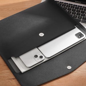 May include: A black leather document holder with a magnetic clasp. Two silver smartphones are inside. The holder is open, showing the phones and part of a laptop keyboard on a wooden surface.