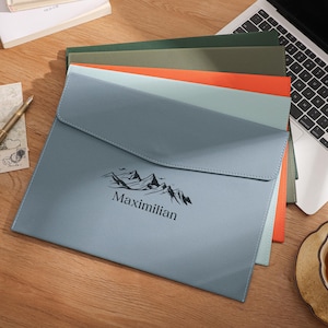May include: A stack of document holders in various colors, including blue, green, and orange, with a laptop and a cup of tea on a wooden surface. The front document holder is light blue and has the name "Maximilian" and a mountain graphic.