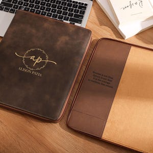 Pode incluir: Duas pastas de documentos em couro castanho. Uma pasta fechada exibe um monograma dourado e o nome "ALISON PATIA". A outra está aberta, revelando um interior bege e texto impresso. Um laptop e livros estão no fundo.