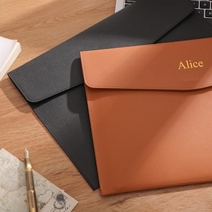 May include: Two leather document holders, one black and one tan, are displayed on a wooden surface. The tan holder has the name "Alice" embossed in gold. A pen, papers, and a laptop keyboard are also visible.