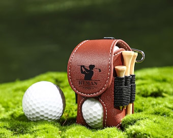 May include: A brown leather golf ball and tee holder with the name "Hyman" printed on it. The holder has a golfer silhouette and is sitting on green moss with two golf balls.