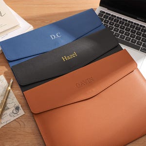 May include: Three personalised leather document holders in blue, black, and brown, each with a name or initials embossed on the front. A laptop, book, pen, and stationery are also visible.