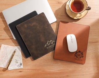 Op de afbeelding: Een laptop, drie leren muismatten, een witte muis, een pen en een theekopje op een houten bureau. De muismatten zijn bruin, zwart en donkerbruin. De donkerbruine mat heeft een bergafbeelding en de tekst "WASHAKI". De bruine mat heeft een wapenafbeelding en de tekst "WEAYAYA".