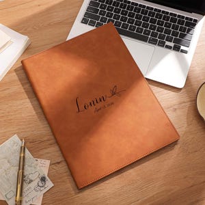 May include: A tan leather portfolio with the name "Lonin" and the date "April 13, 2026" printed on the front. The portfolio is on a wooden desk next to a laptop, a cup of tea, and stationary.