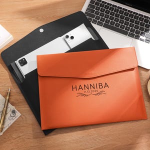 May include: An orange leather document case with the name "HANNIBAL" and the date "11.12.2025" printed on the front. A black leather case is open, revealing a white tablet. A laptop, pen, and teacup are also on the wooden surface.