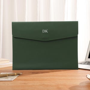 May include: A forest green document case with a flap, displaying the white initials "DK". It sits on a wooden desk, alongside a laptop, a pen, and a cup of tea.